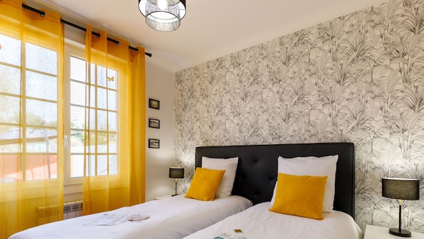 Une chambre d’hôtel avec deux lits, une fenêtre avec des rideaux jaunes, un mur recouvert d’un papier peint à motifs floraux et une table de chevet avec une lampe.