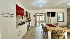 A modern kitchen with a dining area, featuring a wooden table, chairs, and a view through a glass door to an outdoor patio. - Le Corti Sarde Beach Deluxe (Bari Sardo)