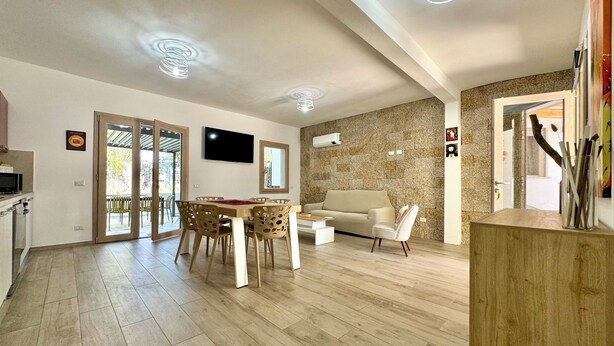 A modern living room with a sofa, dining table, and chairs, a flat-screen TV, and a stone accent wall. - Le Corti Sarde Beach Deluxe (Bari Sardo)