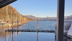 Vue depuis une cabane dotée d’une terrasse en bois, donnant sur une marina avec des bateaux et un paysage montagneux en arrière-plan.