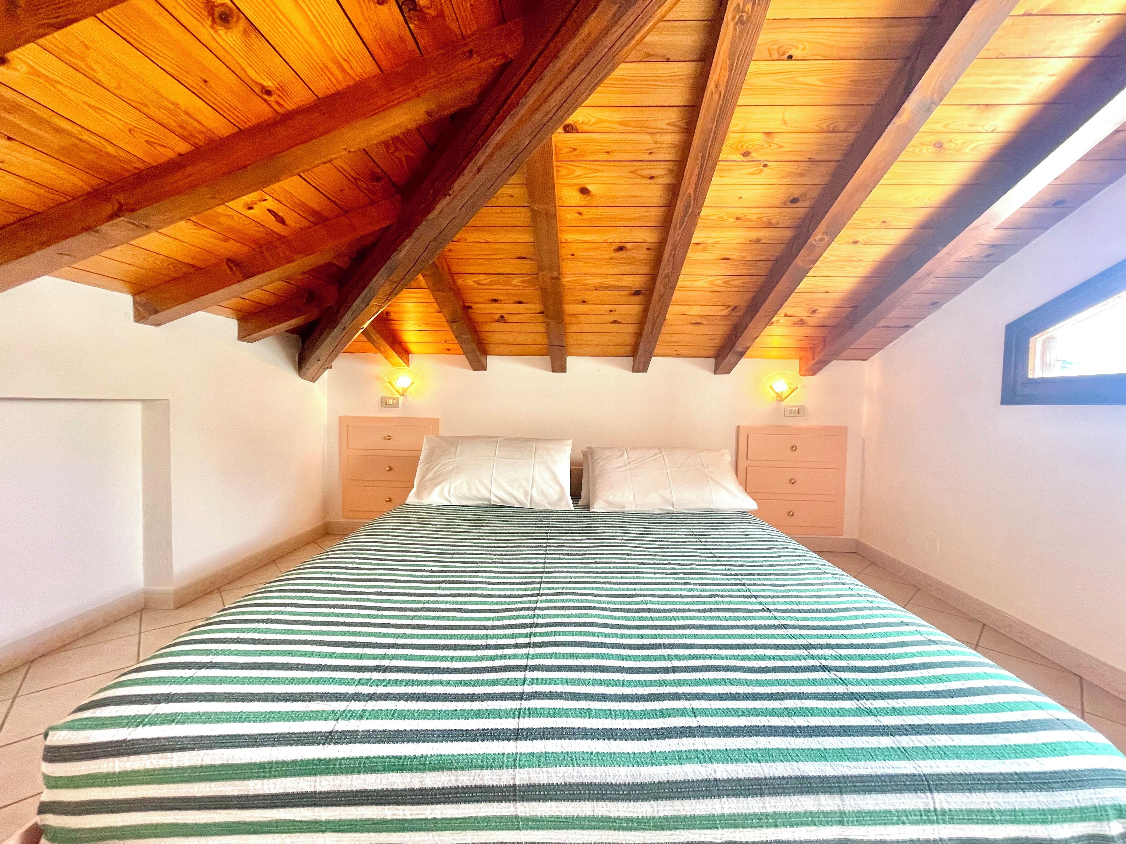 A bedroom with a sloped wooden ceiling, a bed with striped bedding, two bedside tables, and a window on the right wall.