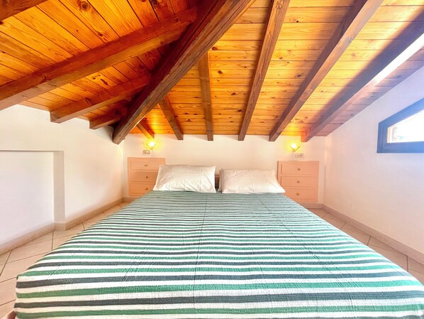 A bedroom with a sloped wooden ceiling, a bed with striped bedding, two bedside tables, and a window on the right wall.
