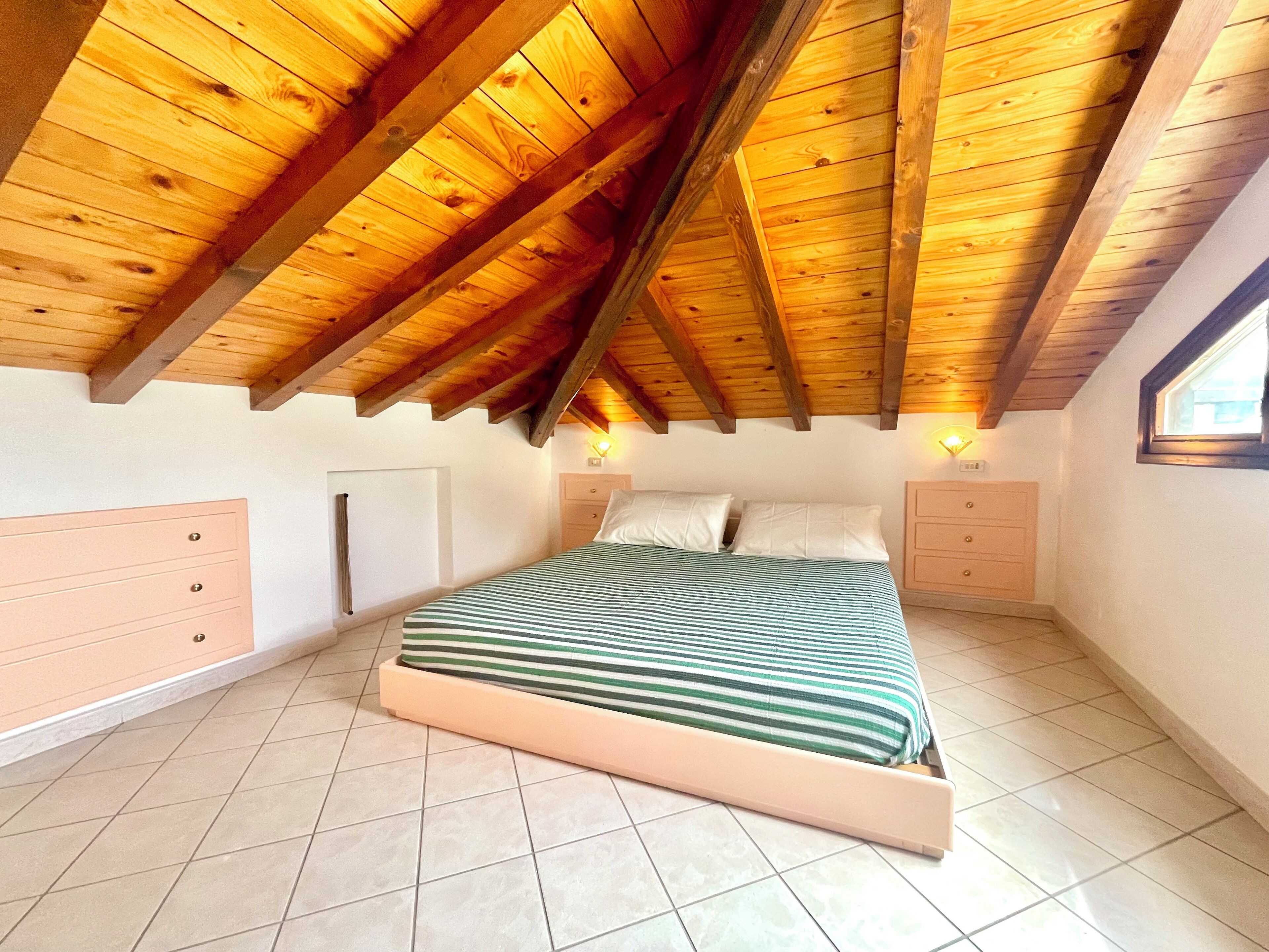 Un dormitorio con techo de madera inclinado, una cama con colcha a rayas, dos mesitas de noche y una ventana en la pared derecha.