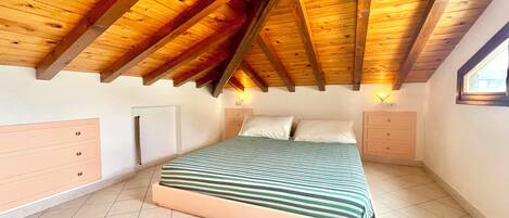 Un dormitorio con techo de madera inclinado, una cama con colcha a rayas, dos mesitas de noche y una ventana en la pared derecha.