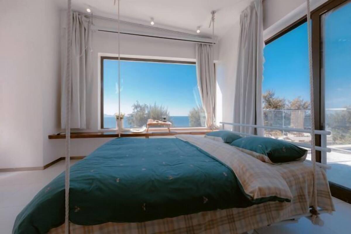 A bedroom with a large window, a bed with a green and beige bedspread, and a view of the outdoors.