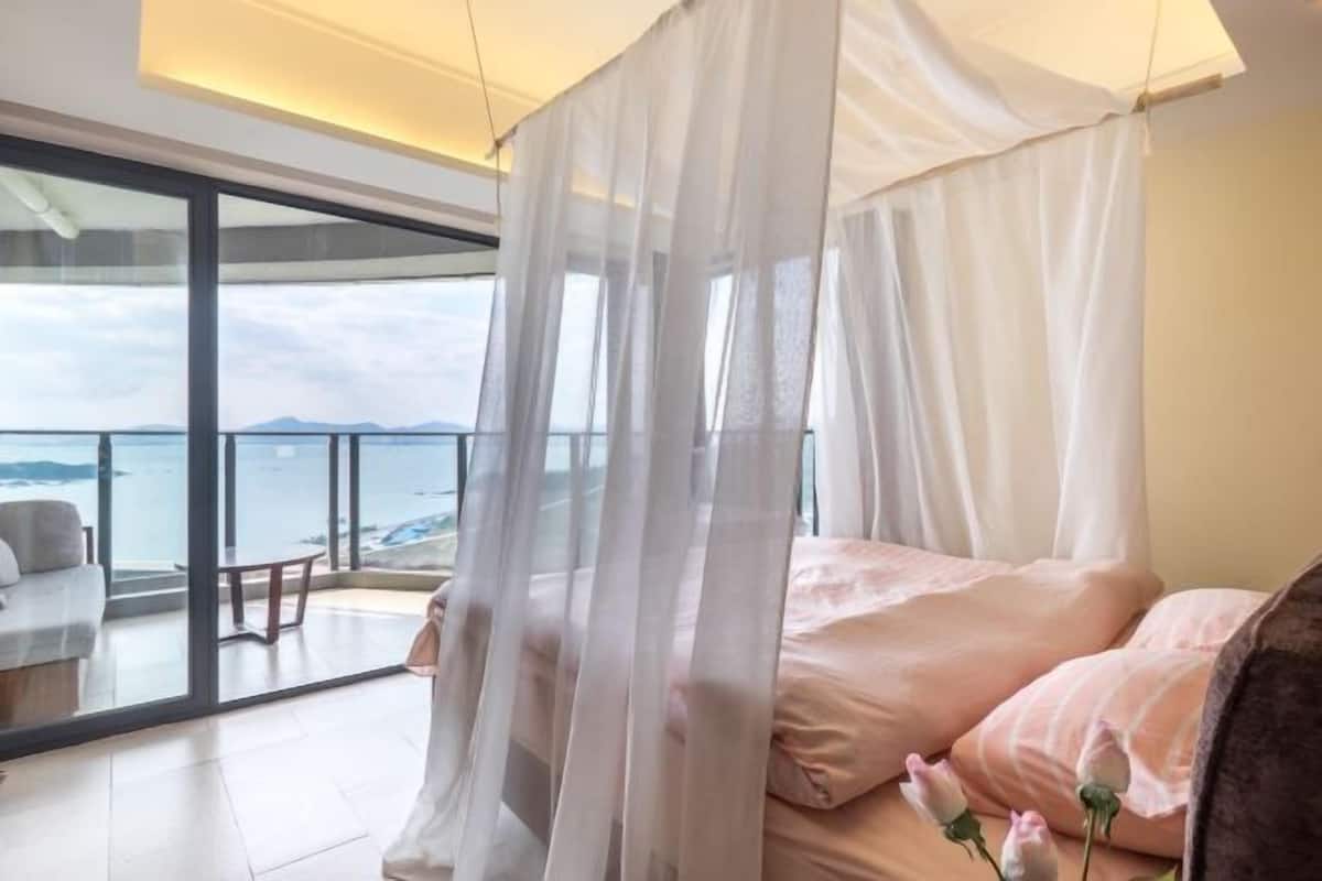A bedroom with a bed, a canopy, a view of the sea, and a balcony with a table and chairs.