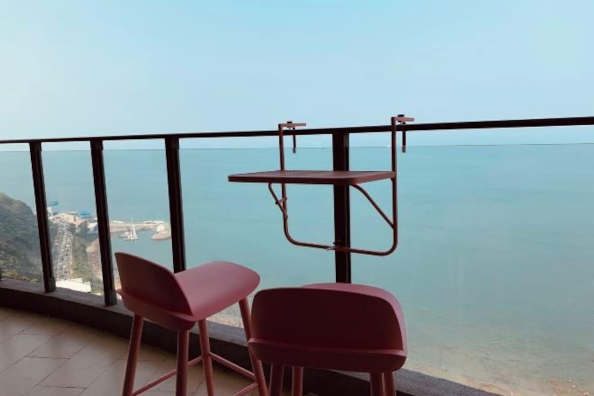 A balcony with two red chairs and a small table, overlooking a coastal view.
