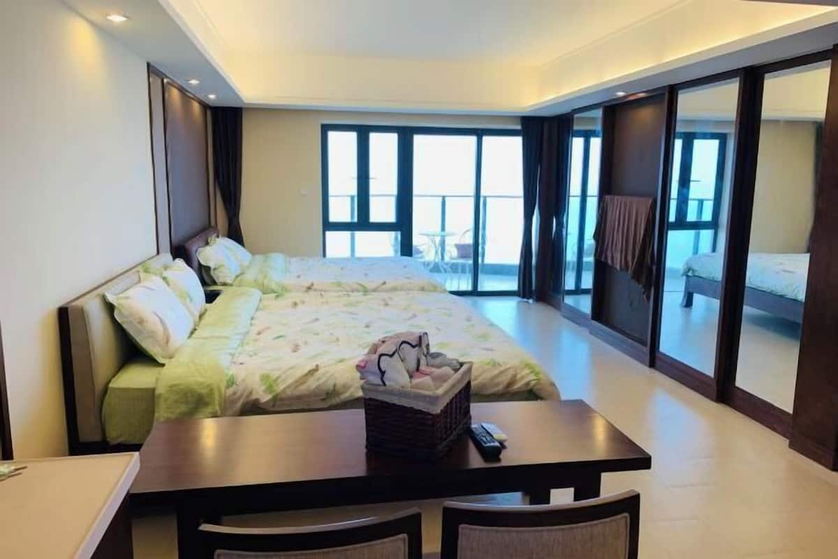 A hotel room with a large bed, a dining table with chairs, a small table with a basket, and a balcony with a chair.