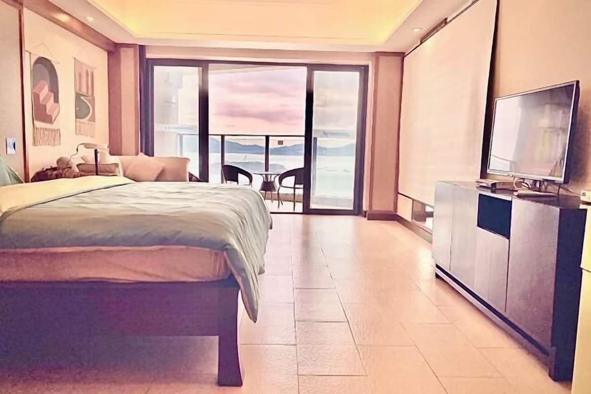 A hotel room with a bed, a TV, a balcony with a table and chairs, and a view of the sea.