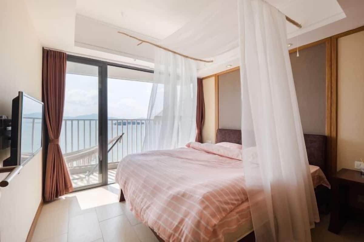 A hotel room with a bed, a TV, a balcony with curtains, and a view of the sea.