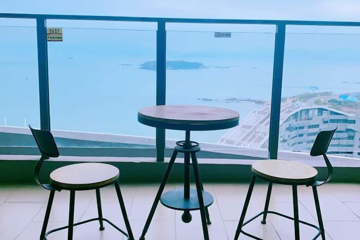 A balcony with two chairs and a small table, overlooking a coastal area with buildings and a bridge.