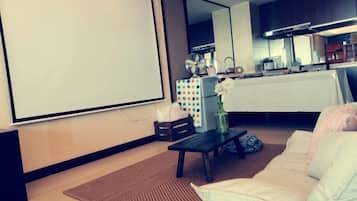 Un salon équipé d’un écran de projection, d’un canapé, d’une table basse et d’un coin cuisine à l’arrière-plan.