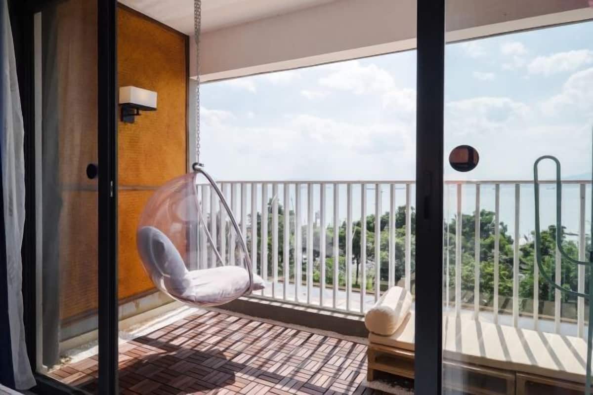 A balcony with a hanging chair, a cushioned bench, and a view of greenery and buildings.