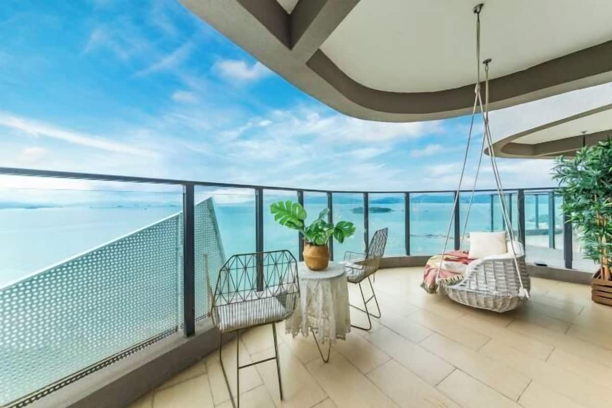 A balcony with a view of the sea, a hanging chair, a small table, and a potted plant.