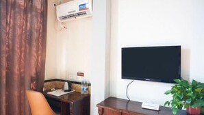 A hotel room with a wooden desk, a chair, a flat-screen TV mounted on the wall, a potted plant, and an air conditioning unit.