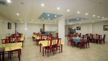 A spacious dining area with multiple tables and chairs, a kitchen in the background, and a tiled floor.