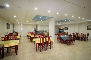 A spacious dining area with multiple tables and chairs, a kitchen in the background, and a tiled floor. - Longdu Business Hotel (Zhaoyuan)