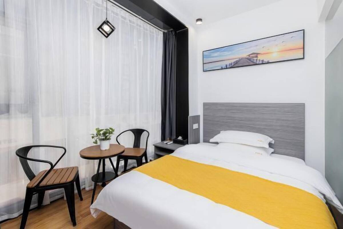 Ein Hotelzimmer mit einem Bett, einem kleinen Tisch mit Stühlen, einem Blumentopf, einem Nachttisch und einem Bild an der Wand.