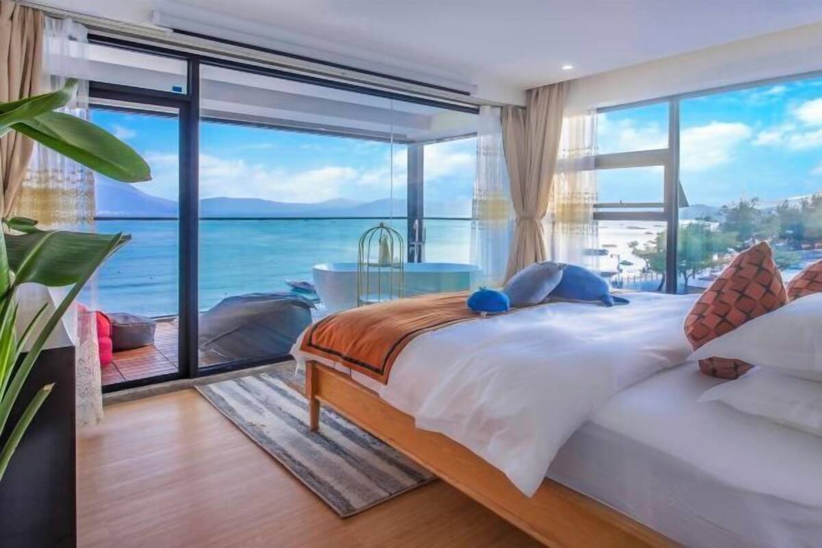 A bedroom with a large window, a bed, a chair, and a view of the sea.