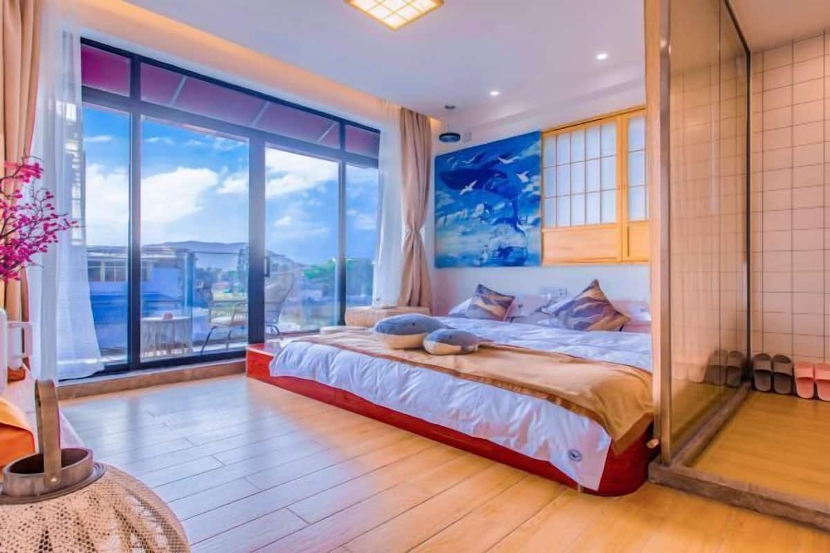 A spacious bedroom with a large bed, a view through floor-to-ceiling windows, and a painting on the wall.