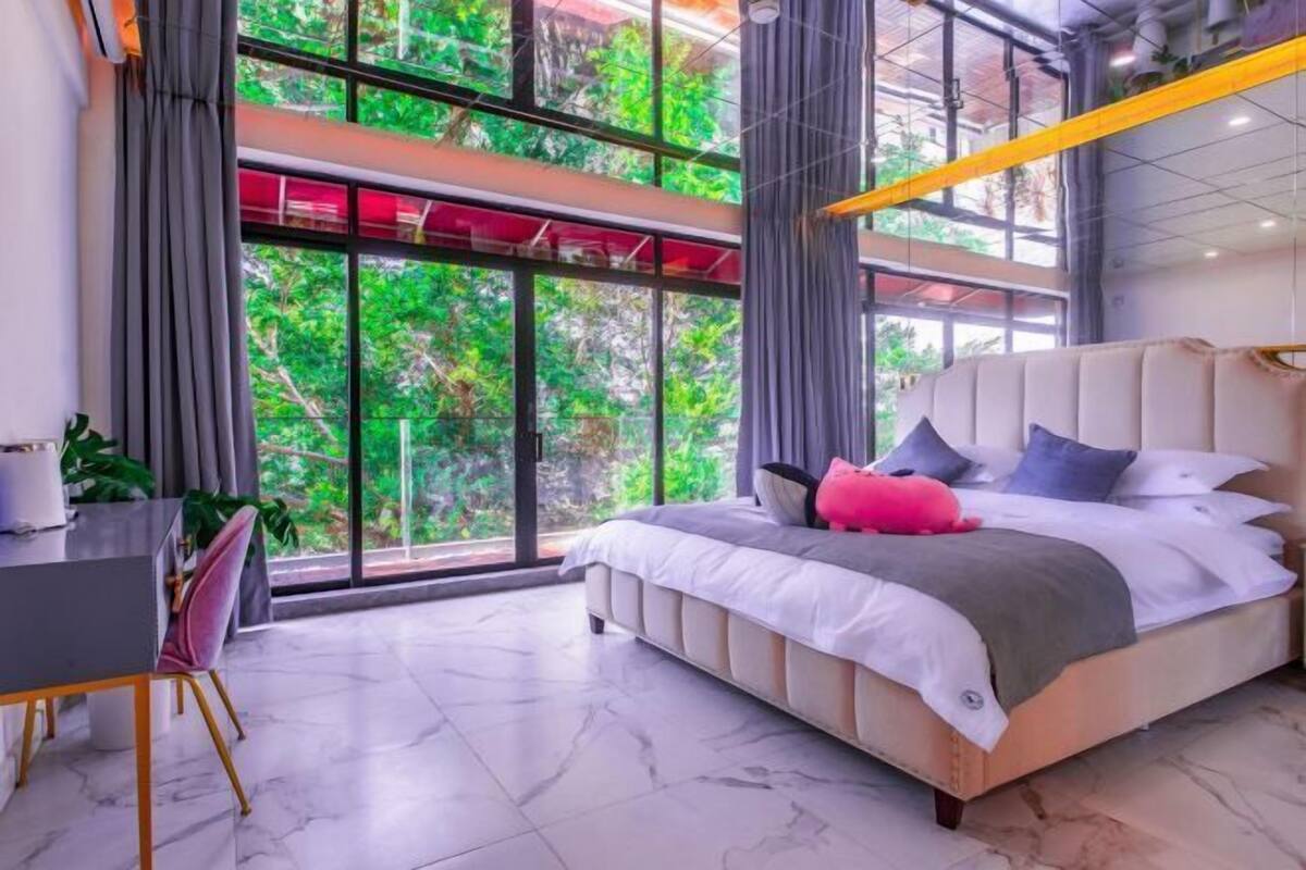 A modern bedroom with a large bed, a desk, and a view of greenery through large windows.