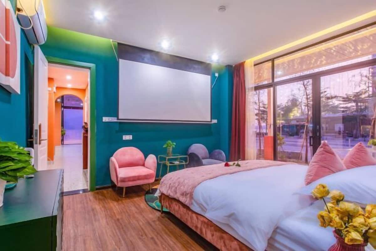 A bedroom with a large bed, a TV, a pink chair, and a window with a view.