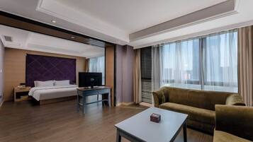 A hotel room with a bed, a TV, a sofa, a coffee table, and large windows.