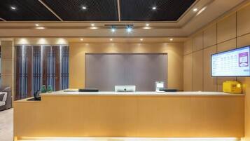 A reception desk with a computer and a monitor, a ceiling with recessed lighting, and a wall-mounted screen displaying information.