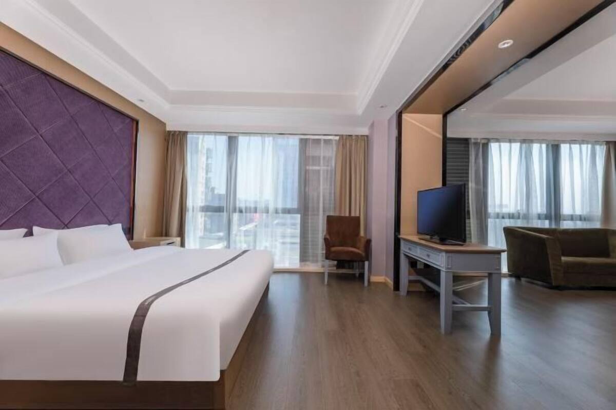 A hotel room with a large bed, a desk, a chair, a television, and a view of the city through large windows.
