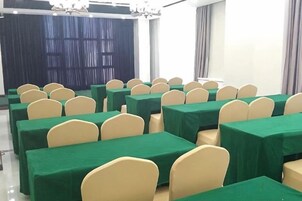 Un salón de conferencias con filas de mesas y sillas, un escenario con cortinas y ventanas amplias.