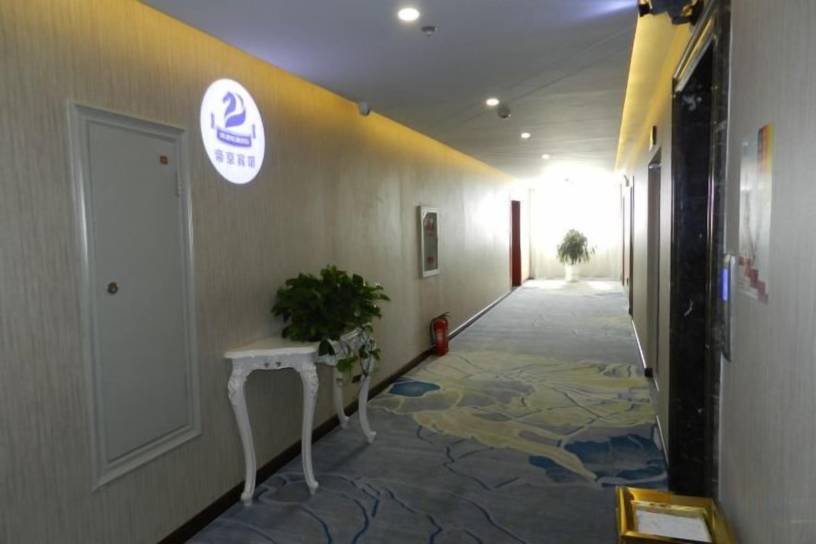 A hotel corridor with a carpeted floor, a white door, a small table with a plant, and a sign with a number.