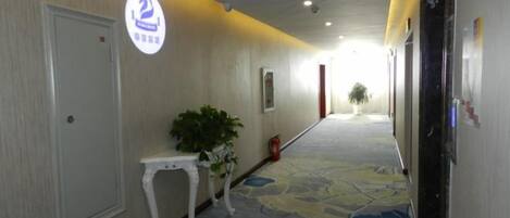 A hotel corridor with a carpeted floor, a white door, a small table with a plant, and a sign with a number.