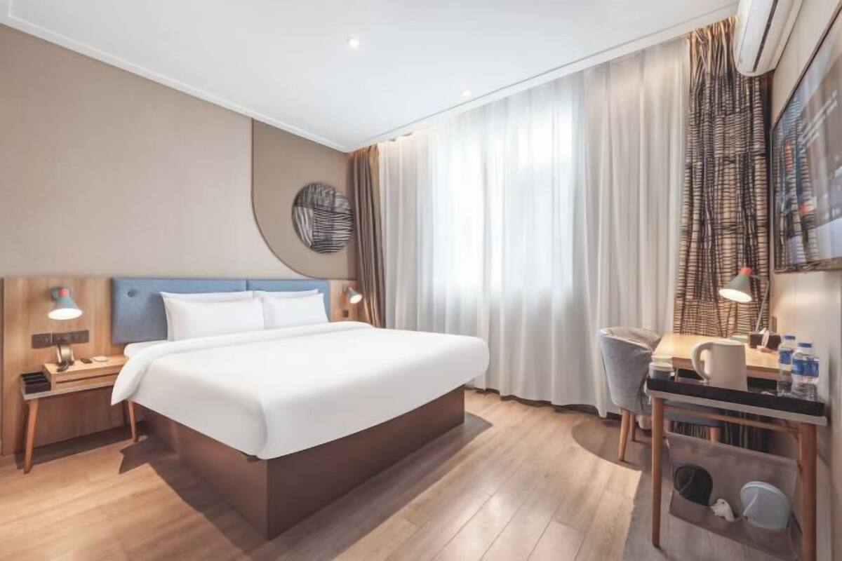 A hotel room with a large bed, bedside tables with lamps, a desk with a chair, and a window with curtains.