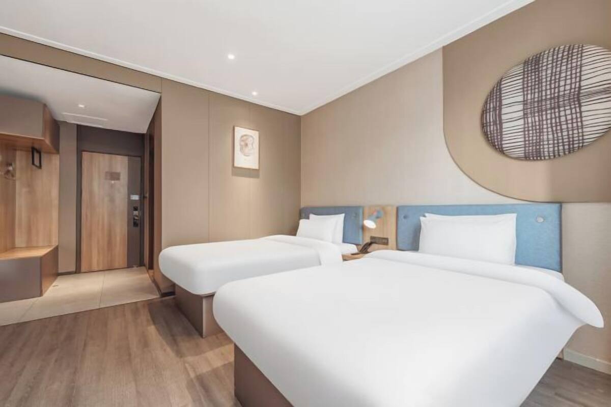 A hotel room with two beds, a wooden wardrobe, a wall-mounted artwork, and a recessed ceiling light.