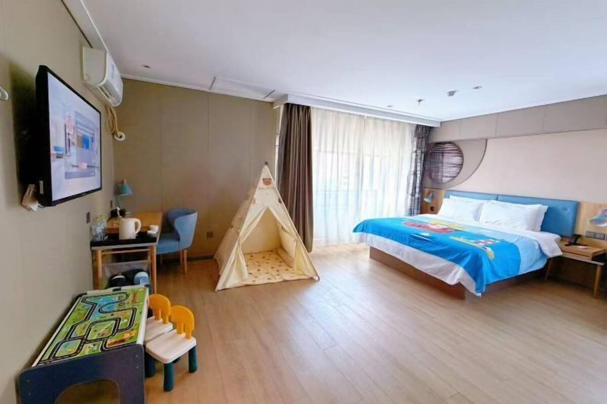 A hotel room with a bed, a desk with a chair, a small table with a chair, a tent, and a wall-mounted TV.