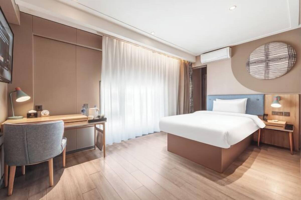 A modern hotel room with a bed, desk, chair, and a large window with curtains.