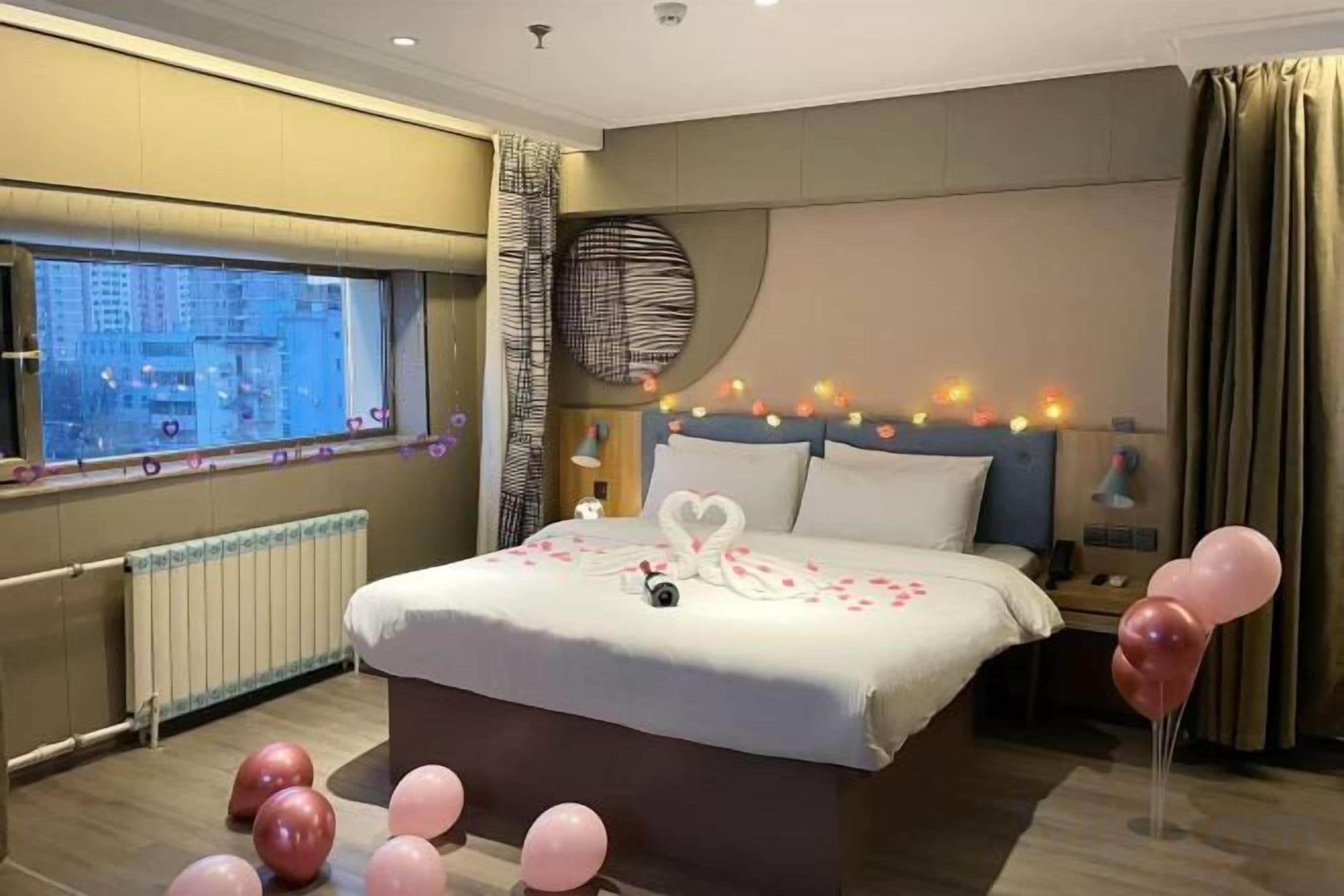 A hotel room with a large bed, a view of the city through a window, and a bedside table with a lamp.