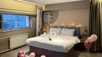 A hotel room with a large bed, a view of the city through a window, and a bedside table with a lamp.