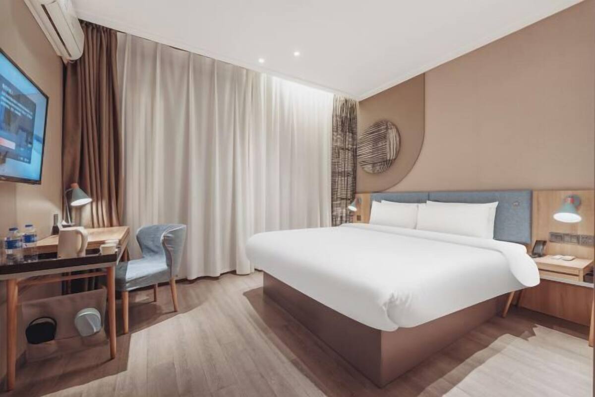 A hotel room with a large bed, a desk with a chair, a TV, and a wall-mounted air conditioner.