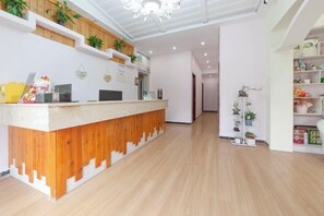 Zona de recepción con mostrador de madera, paredes blancas y una araña. Hay estantes con plantas y una puerta que da a otra habitación.
