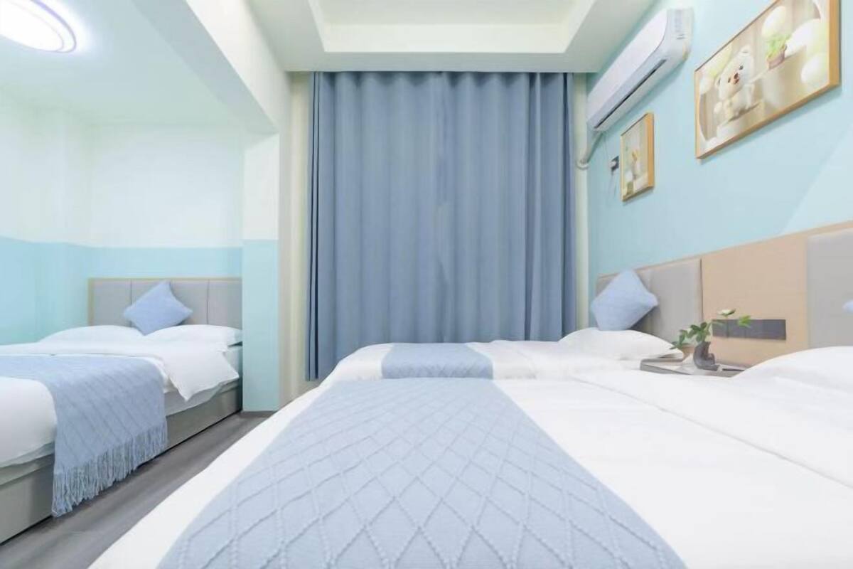 Habitación de hotel con dos camas, un ventanal con cortinas, un aire acondicionado de pared y una pequeña mesa con una planta.