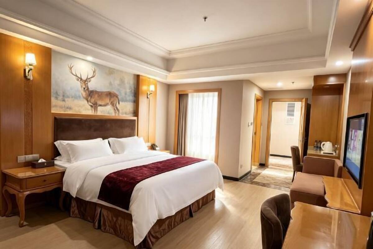 A hotel room with a large bed, a desk, a chair, a painting of a deer, and a television.