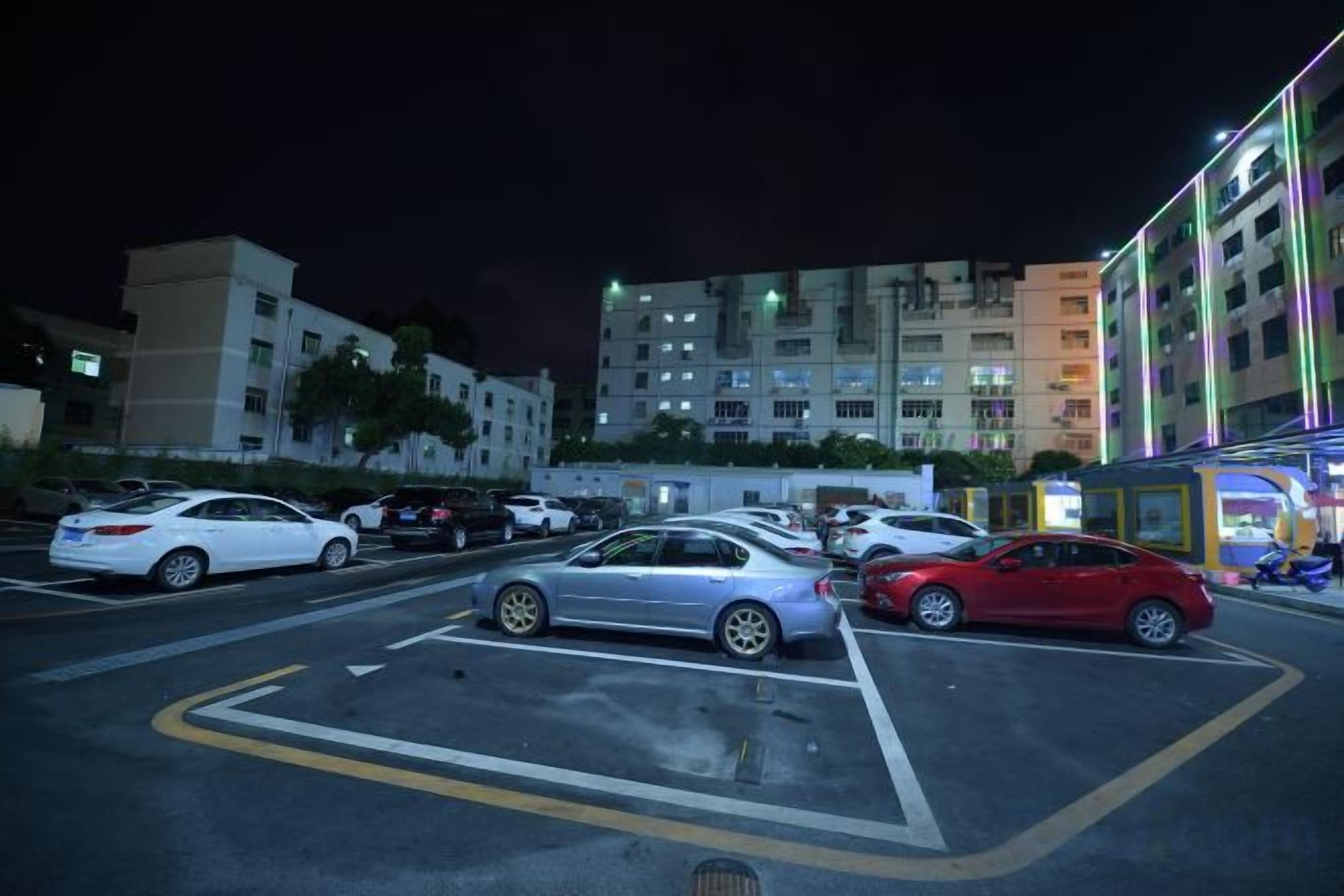 A parking lot with cars and a building with illuminated windows in the background.