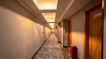 A hotel corridor with a patterned carpet, doors on the right, and a red suitcase on the floor.