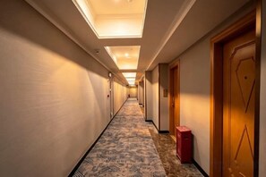 A hotel corridor with a patterned carpet, doors on the right, and a red suitcase on the floor.