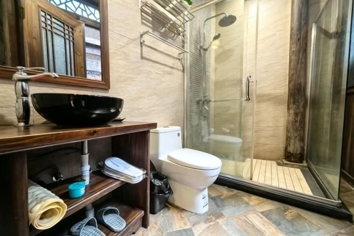 Un baño con un lavamanos tipo recipiente negro, un inodoro, una ducha con puerta de vidrio y un mueble de tocador de madera.