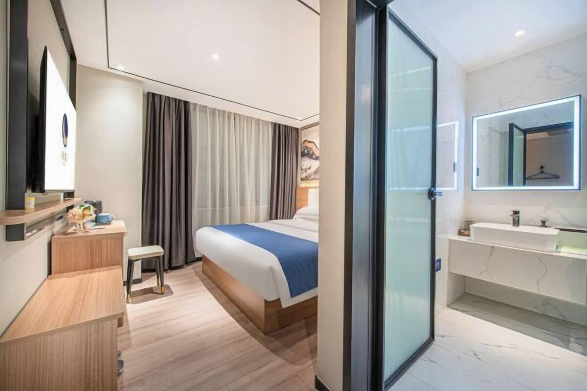 A modern hotel room with a bed, bedside table, chair, and a bathroom with a mirror and sink.