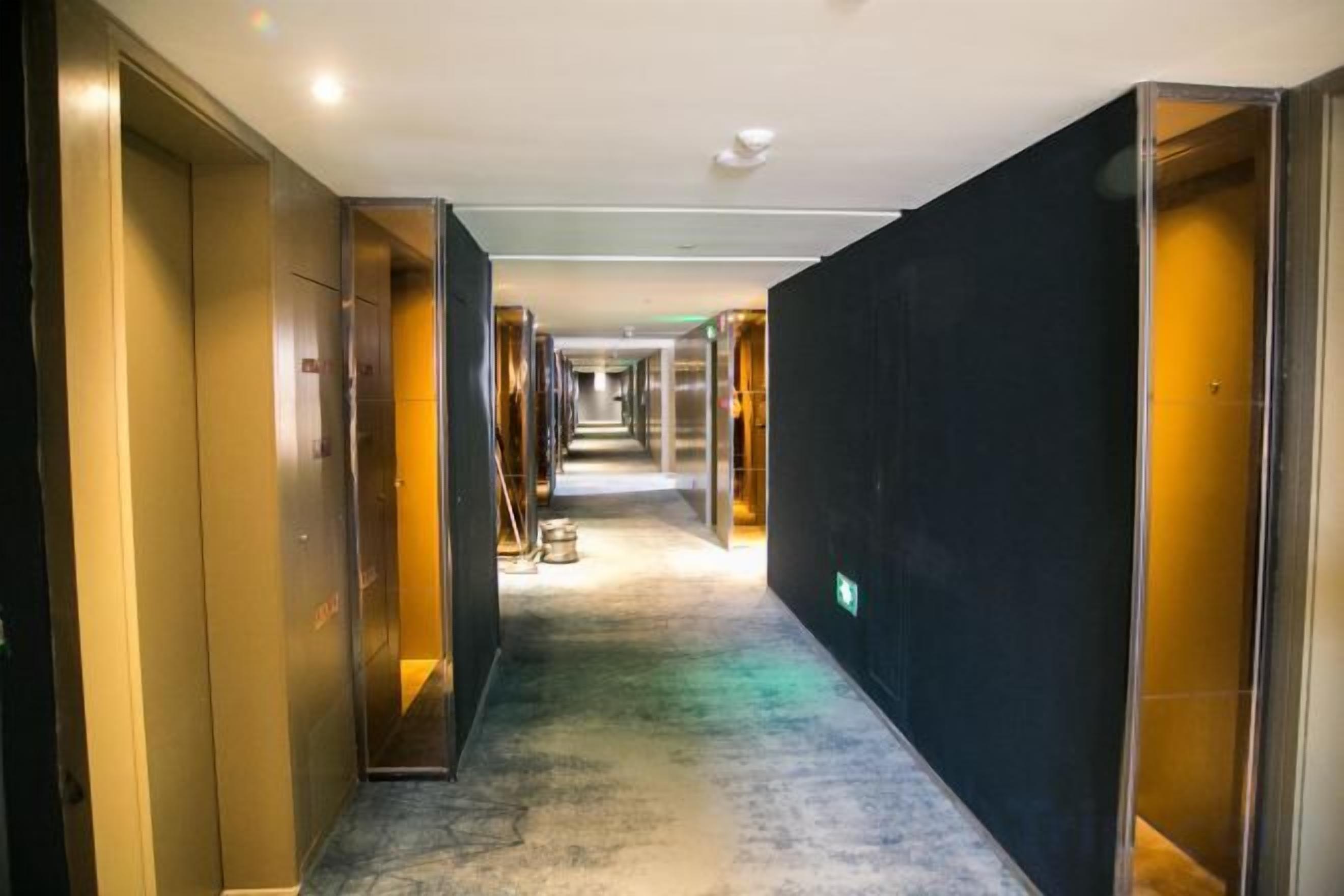A hotel corridor with a carpeted floor, doors on both sides, and a visible emergency exit sign.