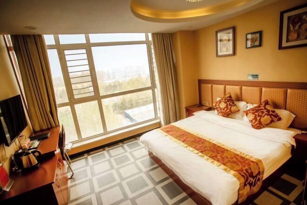 Habitación de hotel con un ventanal grande, cama con ropa de cama estampada, escritorio con ordenador y televisor.