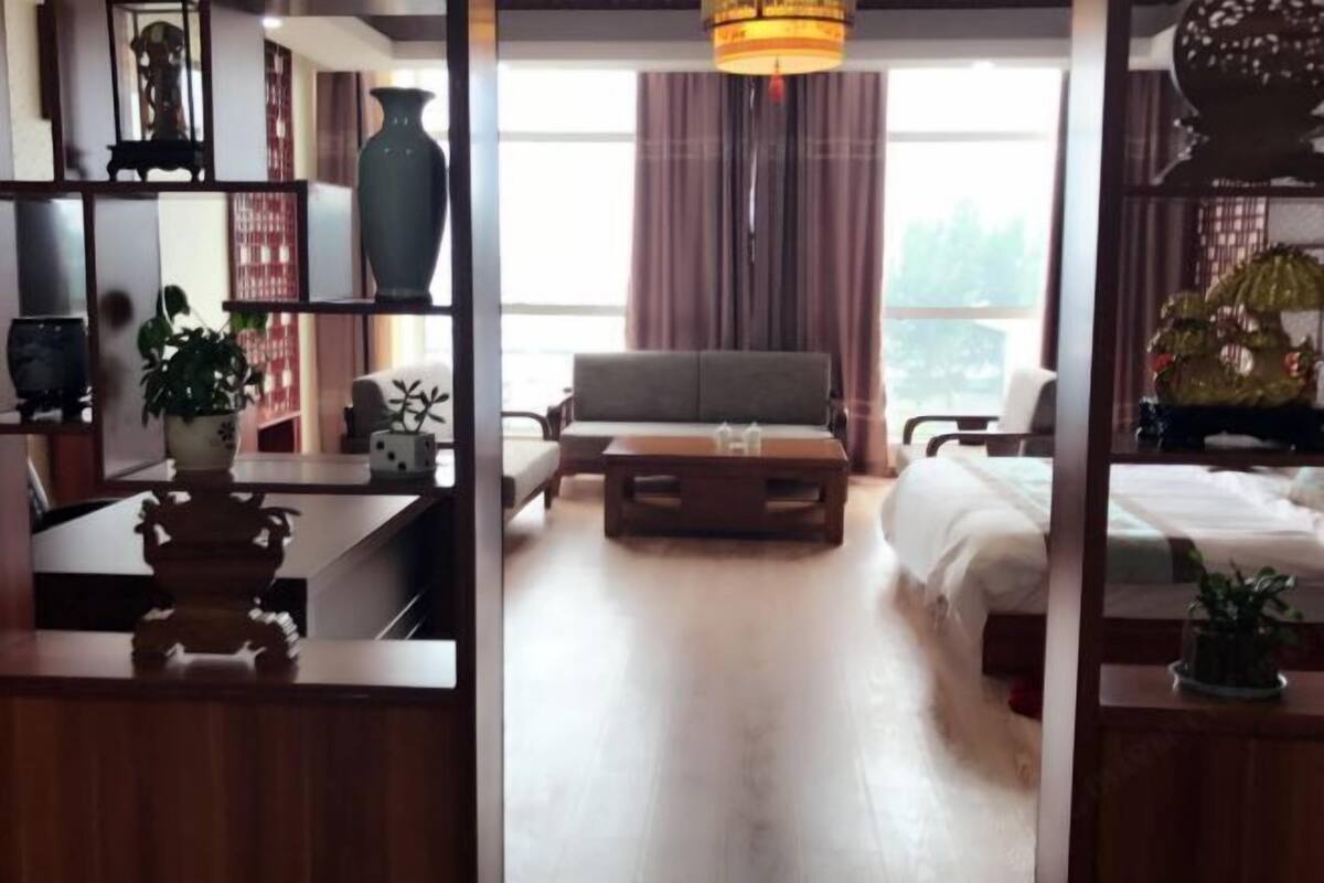 Una habitación con sofá, una mesa de centro y cama. Hay una ventana con cortinas, una planta y un jarrón decorativo.